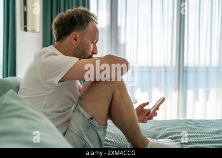 Ein Mann in lässiger Kleidung sitzt auf dem Bett und blickt in einem hell beleuchteten Raum auf sein Smartphone. Stockfoto