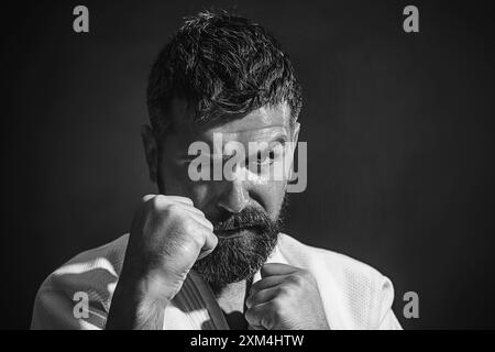 Karate-Mann in Kampfposition. Bärtiger Karate-Mann in weißem Kimono für Sambo, Judo, Jujitsu mit Händen in Fäusten, bereit zum Kampf. Gemischtes Kriegswesen Stockfoto