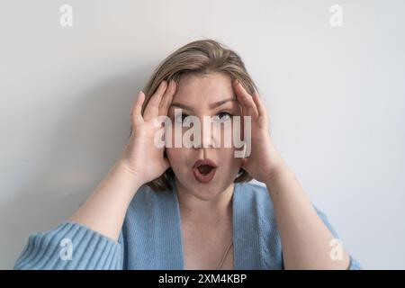 Eine junge Frau in Übergröße sieht überrascht aus, mit offenem Mund und Händen auf dem Kopf. Ausdruck von Schock und Staunen, Porträt mit Blick auf die Kamera. Stockfoto