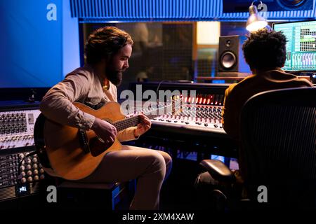 Sänger kreiert einen neuen Hit im Studio mit Gitarrenlauf und arbeitet mit Audioexperten zusammen, um Musik zu produzieren. Songwriter singt eine Schallplatte auf elektroakustischen Instrumenten im Kontrollraum mit Soundboard. Stockfoto