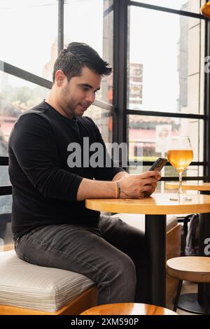Junger Latino-Mann, der an einem Bartisch sitzt und sein Handy benutzt, während er ein Glas Craft-Bier genießt. Die entspannte Atmosphäre und die moderne Bar sind hoch Stockfoto