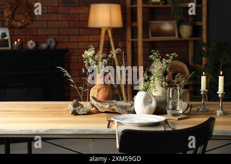 Set aus sauberem Geschirr, Vasen mit schönen Blumen, Steinen und brennenden Kerzen auf Holztisch im stilvollen Esszimmer Stockfoto