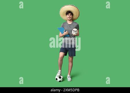 Junger männlicher Fußballspieler mit Sombrero, bemaltem menschlichen Schädel, Fußball und Klemmbrett auf grünem Hintergrund Stockfoto