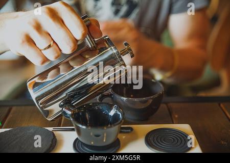 Ein Mann gießt ein Getränk Kaffee oder Tee aus einem Metallkessel in schöne schwarze Tassen in einem gemütlichen, verschwommenen Innenraum Stockfoto