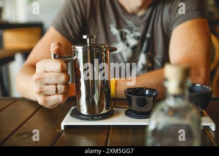 Ein Mann gießt ein Getränk Kaffee oder Tee aus einem Metallkessel in schöne schwarze Tassen in einem gemütlichen, verschwommenen Innenraum Stockfoto