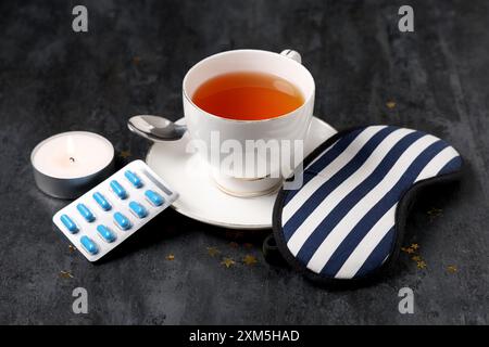 Tasse Tee mit Schlafmaske, Pillen und brennender Kerze auf schwarzem Hintergrund Stockfoto