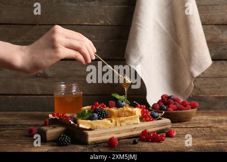 Frau, die mit Honig und Brett süßer belgischer Waffeln mit verschiedenen frischen Beeren auf hölzernem Hintergrund einen Dipper hält Stockfoto