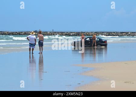 Ein Strandgänger, der sein Auto am Meeresrand parkte, fand sich feststecken; zwei Frauen in String-Bikinis bieten an, ihn mit ihrem Fahrzeug und ihrer Winde herauszuziehen. Stockfoto