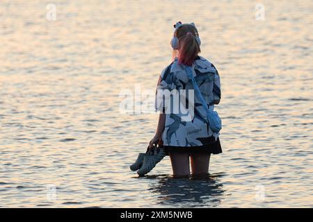 St. Petersburg, Russland. Juli 2024. Ein Mädchen hält Sandalen und hört Musik, während er im Golf von Finnland steht. (Foto von Artem Priakhin/SOPA Images/SIPA USA) Credit: SIPA USA/Alamy Live News Stockfoto