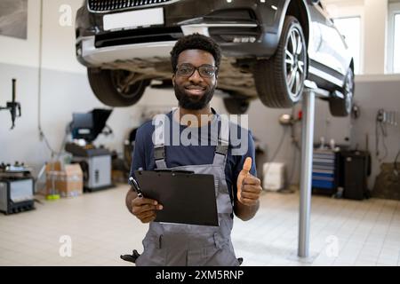 Automechaniker lächelnd mit Daumen nach oben und Klemmbrett in der Werkstatt Stockfoto