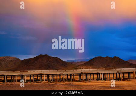 Lebendiger Regenbogen vor dramatischer Kulisse mit gemischtem Himmel, Bögen über dem erhöhten Highway, der sich durch die wunderschöne kalifornische Wüste schlängelt, raues Gelände und vib Stockfoto