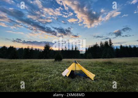 Gelbes Zelt auf grasbewachsenem Feld in der Abenddämmerung, mit atemberaubendem Himmel voller farbenfroher Wolken am Abend. Der Sonnenuntergang strahlt warm aus und schafft eine ruhige und malerische Umgebung, umgeben von immergrünen Bäumen. Stockfoto