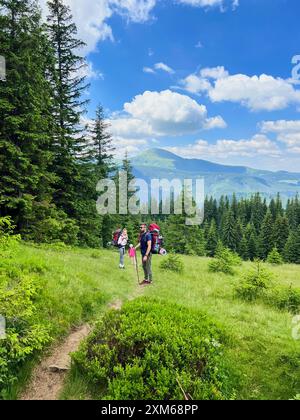 Eine dreiköpfige Familie, zwei Erwachsene mit großen Rucksäcken und ein Kind in rosa Jacke und Hut, hält auf einem grasbewachsenen Hügel. Atemberaubende Berglandschaft mit üppigen grünen Bäumen und klarem blauem Himmel im Hintergrund. Stockfoto