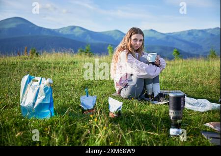 Die Touristin sitzt auf einem grasbewachsenen Hügel und hält den Becher. Reisender mit langen blonden Haaren. In der Nähe tragbarer Campingkocher, Essensbeutel, blaue Tasche. Die malerische Bergkulisse sorgt für eine ruhige Atmosphäre im Freien. Stockfoto