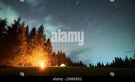 Das Lagerfeuer leuchtet warm neben mehreren beleuchteten Zelten auf einer grasbewachsenen Rodung unter sternenübersätem Himmel mit verstreuten Wolken. Hohe Kiefern bilden die Szene und betonen die ruhige Schönheit der Nacht in der Natur. Stockfoto