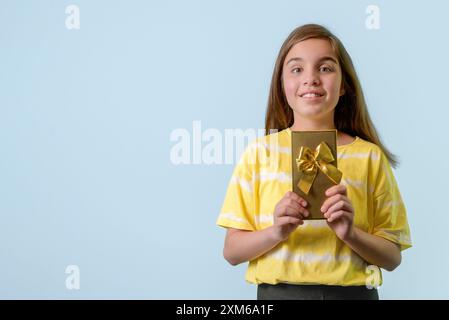 Ein fröhliches Mädchen im Teenageralter hält ein Geschenk in einem vergoldeten Paket mit einer Schleife in den Händen. Hellblauer Hintergrund Stockfoto