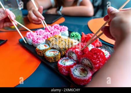 Sushi-Brötchen und Leute mit chinesischen Stäbchen in der Hand nehmen Sushi. Japanisches Restaurant, schwarzer Teller mit verschiedenen gebackenen Brötchen und Philadelphia Stockfoto