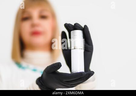 Kosmetikerin, Schönheitstherapeutin Frau in einem weißen Labormantel hält eine weiße Flasche Hautpflegemittel. Sie trägt schwarze Handschuhe. Stockfoto