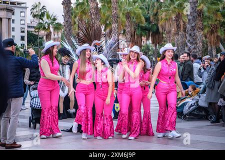 Farbenfrohe Gruppe Barbie-Frauen in leuchtend rosa Outfits mit weißen Cowboyhüten bei einer Veranstaltung im Freien, mit Palmen gesäumten Hintergrund. Stockfoto