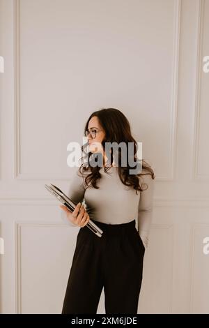 Hispanische Frau mit Notizbüchern und Laptop in der Hand, weiße getäfelte Wand Stockfoto