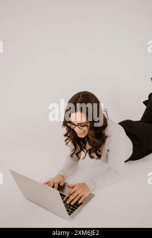 Die junge hispanische Frau liegt auf dem Boden, arbeitet an einem Laptop und lächelt Stockfoto