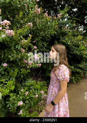 Ein junges Mädchen in einem rosafarbenen Blumenkleid, das Blumen im Garten untersucht. Stockfoto