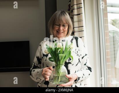 Frau in ihren 70ern lächelt Blumen in einer Vase, die als Geschenk gegeben wurde Stockfoto