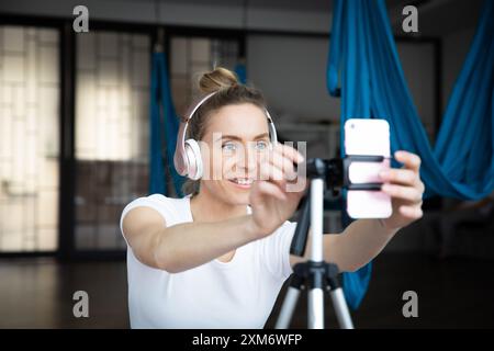 Junge schöne Frau in Kopfhörern lächelt in die Smartphone-Kamera und unterrichtet online Yoga-Kurs Stockfoto