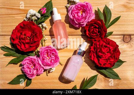 Zwei matte Tropfflaschen mit einem natürlichen Gesichts- und Körperpflegeprodukt auf Holzhintergrund zwischen Rosen. Pflege- und Schönheitskonzept. Naturkosmetik Stockfoto