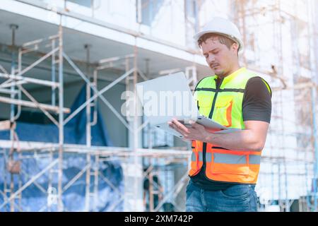 Bauarbeiter, der Sicherheitsausrüstung trägt und auf einer Baustelle einen Laptop verwendet, um Projektdetails zu verwalten. Stockfoto