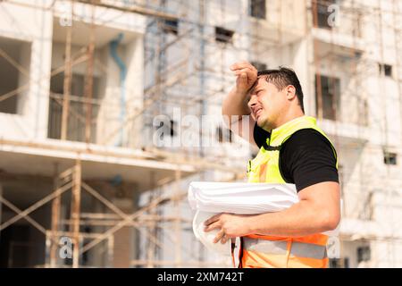 Bauarbeiter, der eine Pause macht und unter der Sonne schwitzt, einen Schutzhelm hält, mit einem Baugerüst im Hintergrund. Stockfoto