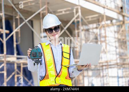 Bauarbeiterinnen, die Sicherheitsausrüstung tragen und einen Laptop verwenden, bedienen Lasermessgeräte auf einer Baustelle. Stockfoto