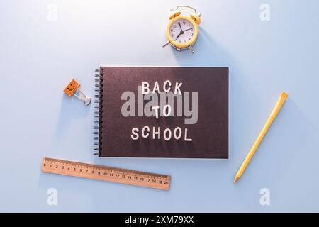 Schwarzes Notizbuch Mit Den Worten „Back To School“ In Weißen Buchstaben Auf Blauer Oberfläche Mit Gelbem Bleistift, Lineal Und Wecker Stockfoto