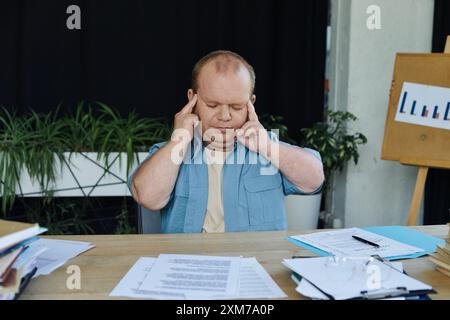 Ein Mann mit Inklusivität sitzt mit geschlossenen Augen an seinem Schreibtisch und nimmt sich einen Moment Zeit, um sich inmitten eines Meeres von Papierkram zu zentrieren. Stockfoto