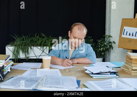 Ein Mann mit Inklusivität sitzt an einem Schreibtisch, schreibt auf einem Blatt Papier, umgeben von Papierkram und einer Tasse Kaffee. Stockfoto