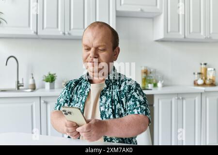 Ein Mann mit Inklusivität, der ein gemustertes Hemd trägt, sitzt an einem Küchentisch und schaut aufmerksam auf sein Smartphone. Stockfoto