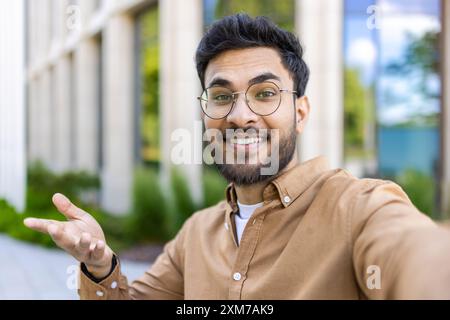 Lächelnder Mann mit Brille, der Selfie im Freien in der Nähe des Bürogebäudes macht, mit Bäumen im Hintergrund. Ein Mann trägt ein lässiges braunes Hemd, wirkt glücklich und selbstbewusst in sonniger Umgebung. Stockfoto