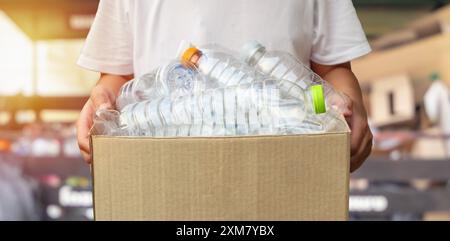 Hand hält braune Recycling-Müllkiste mit Plastikflaschen mit kleinem Recycling-Stationshintergrund Stockfoto