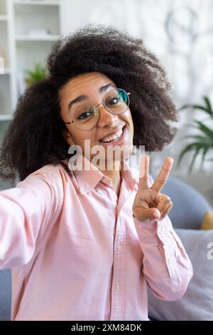 Fröhliche junge Frau mit lockigem Haar und Brille, die Friedenszeichen macht, während sie Selfie im hellen Wohnzimmer macht, mit rosa Hemd. Konzept von Glück, Selbstvertrauen und Positivität moderner Lebensstil Stockfoto