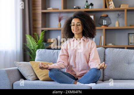 Frau sitzt im Kreuz auf der Couch im Wohnzimmer und meditiert mit geschlossenen Augen. Konzept von Achtsamkeit, Entspannung und Selbstversorgung. Friedliche Inneneinrichtung mit moderner Einrichtung. Stockfoto