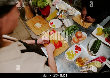 Über der Schulter Schuss von zwei Köchen, die Paprika und Pilze auf hölzernen Schneidebrettern schneiden, Stahl Küchentheke bedeckt mit einer riesigen Anzahl von Tellern mit Zutaten Stockfoto