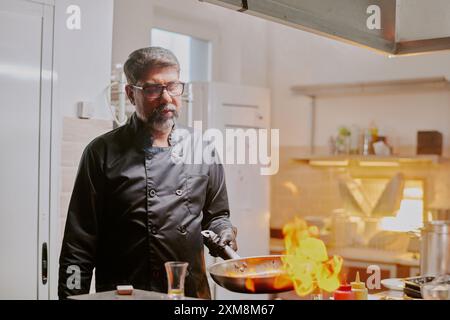 Hochqualifizierter Koch in schwarzer Uniform gekleidet Bratmahlzeit mit Feuer in der modernen Küche des beliebten Restaurants Stockfoto