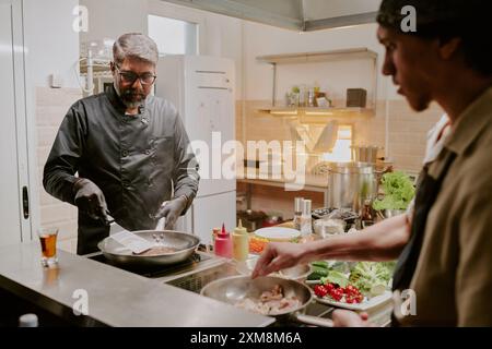 Professioneller indischer Koch in schwarzer Jacke und mit schwarzen Latexhandschuhen, der schwarzes angus-Steak anbrät, während sein Sous Chef Speck frittiert Stockfoto