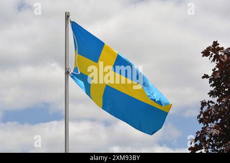 Die Nationalflagge Schwedens fliegt an einem schönen Sommertag mit blauem Himmel und weißen Wolken einen Fahnenmast hoch. Stockfoto