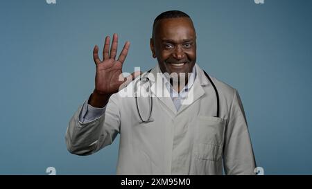 Afroamerikanischer Mann freundlich einladend lächelnder männlicher Arzt medizinischer Therapeut medizinischer Spezialist im blauen Hintergrund Studio, das die Kamera winkt Stockfoto