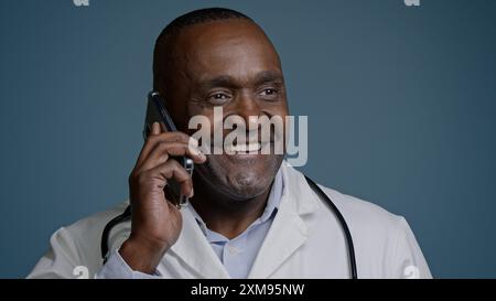 Reifer afroamerikanischer männlicher Arzt Berater spricht auf Handy im blauen Hintergrund Studio Mann Facharzt Arzt in medizinischem Kleid Fernbedienung Stockfoto