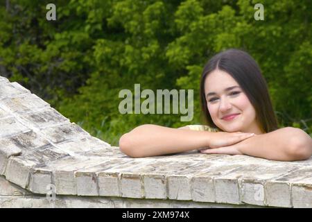 Weibliche Teenager legt ihre Ellbogen auf eine Ziegelwand, während sie hinüberblickt. Sie hat lange Haare und lächelt. Stockfoto