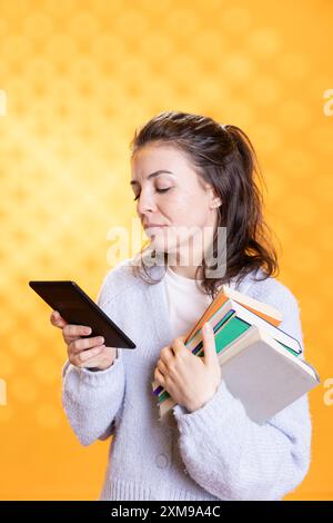 Frau, die eBook Reader und Stapel Bücher hält, isoliert über Studiohintergrund. Person mit einem Stapel von Lehrbüchern im Arm, die wissenschaftliche Arbeiten für Forschungsprojekte auf Tablets liest Stockfoto