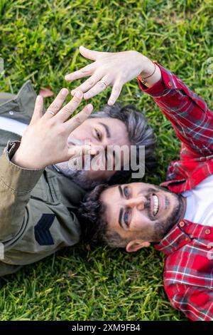 Blick von oben auf ein hispanisches schwules Paar, das auf dem Gras liegt, während es ihre Eheschließungen zeigt Stockfoto