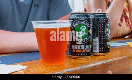 Erfrischendes Bernsteinlager in einem Plastikbecher und zwei ungeöffnete Dosen auf einem Holztisch New York NY USA 30.07.2023 Stockfoto